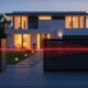 A red infrared beam detectors stretches across the front gate of a modern house at dusk. The beam originates from a sensor on the left gatepost and connects to a sensor on the right, forming a security barrier. The two-story house, with its warm interior lights on, is surrounded by a well-maintained garden and a paved walkway leading to the front door. The sky in the background transitions from deep blue to orange, with the setting sun casting a warm glow on the scene.